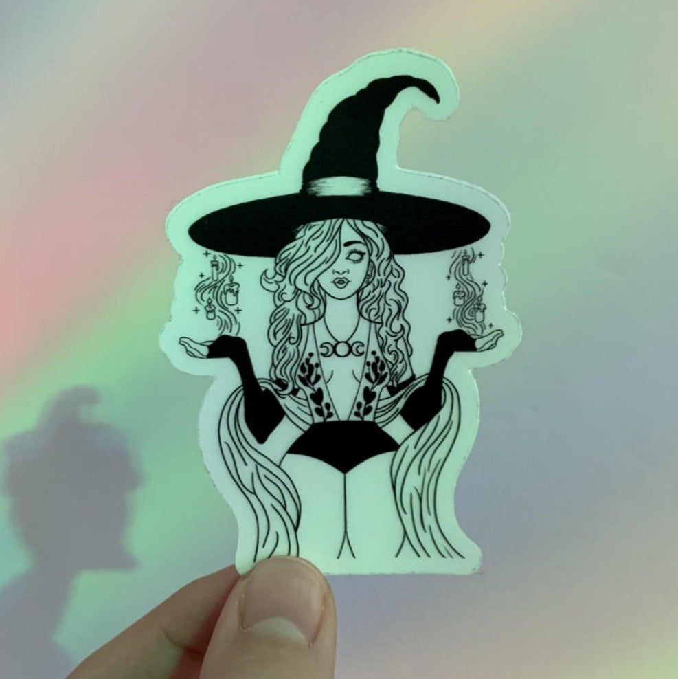 Hedge Witch Sticker design features a lovely woman wearing a deep cut mesh bodysuit with leaves over the top, long black gloves, a necklace with the divine feminine goddess symbol and a shawl scarf. She has a large halloween witch hat on and has two sets of melting wax candles floating above her hands with stars and sparkles swirling around them. She is certainly not evil, but she does not have any irises for she stayed in the spirit world for too long. This pic shows the spooky witchy sticker with rainbows